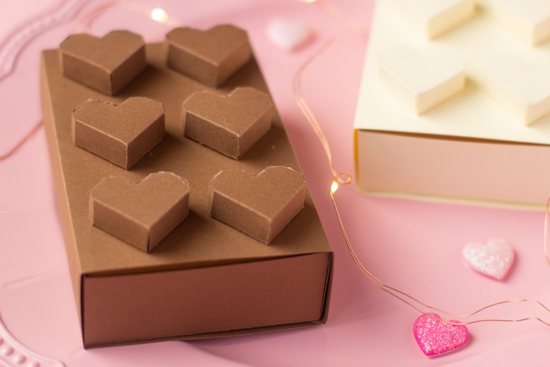 May include: A brown box with six heart-shaped compartments, perfect for holding small gifts or treats. The box is sitting on a pink surface with string lights and pink hearts.