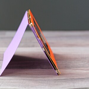 May include: A purple card folded into a triangular shape, standing upright on a wooden surface. The card is decorated with a layered orange and gold design.
