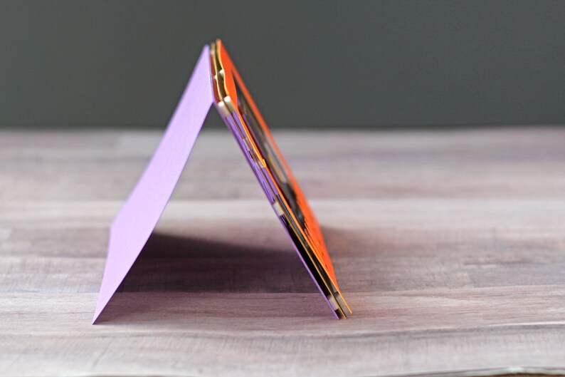 May include: A purple card folded into a triangular shape, standing upright on a wooden surface. The card is decorated with a layered orange and gold design.