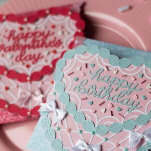 Pu&ograve; includere: Due biglietti d'auguri fatti a mano con sfondo rosa e bianco. La carta a sinistra ha un bordo rosso a forma di cuore con il testo "Buon San Valentino". La carta a destra ha un bordo turchese a forma di cuore con il testo "Buon compleanno".