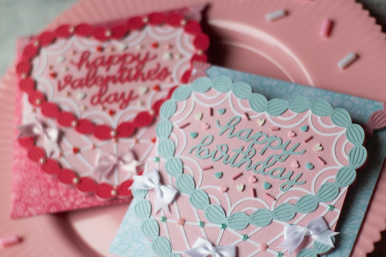 Pu&ograve; includere: Due biglietti d'auguri fatti a mano con sfondo rosa e bianco. La carta a sinistra ha un bordo rosso a forma di cuore con il testo "Buon San Valentino". La carta a destra ha un bordo turchese a forma di cuore con il testo "Buon compleanno".