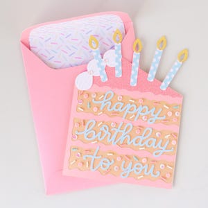 Może przedstawiać: R&oacute;żowa kartka urodzinowa w kształcie ciasta z napisem "happy birthday to you" w niebieskim skrypcie. Karta ma cztery niebiesko-białe świece z ż&oacute;łtymi płomieniami. Karta znajduje się w r&oacute;żowej kopercie z wzorzystą wkładką.