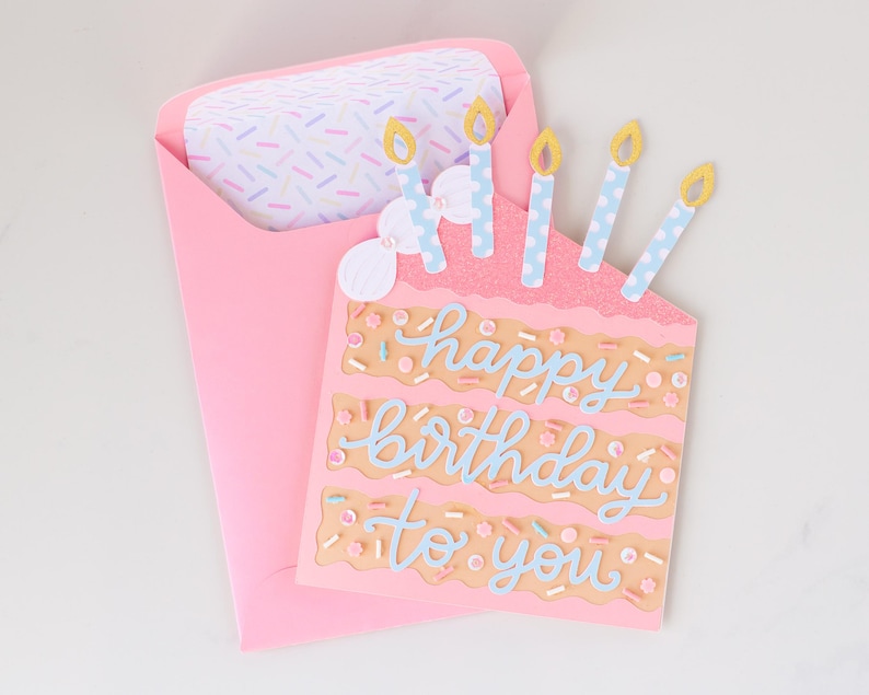 Może przedstawiać: R&oacute;żowa kartka urodzinowa w kształcie ciasta z napisem "happy birthday to you" w niebieskim skrypcie. Karta ma cztery niebiesko-białe świece z ż&oacute;łtymi płomieniami. Karta znajduje się w r&oacute;żowej kopercie z wzorzystą wkładką.