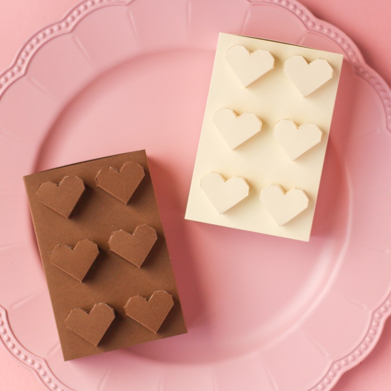 May include: Two boxes of chocolate hearts, one brown and one white, on a pink plate. The boxes are made of cardboard and have a simple design. Each box has six hearts arranged in a grid pattern.
