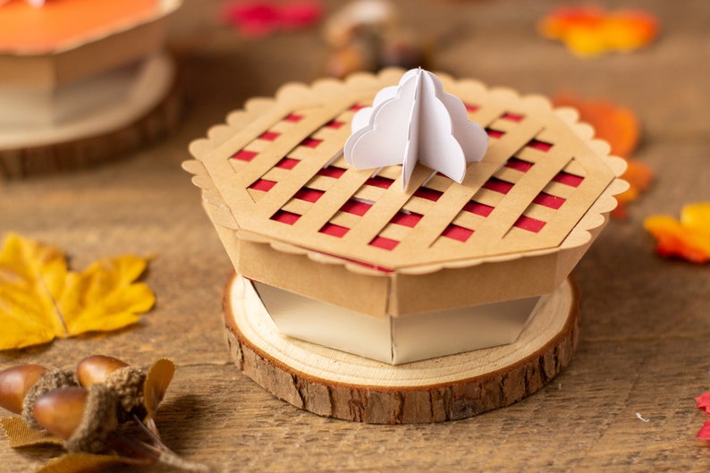 May include: A brown paper pie box with a red and white lattice top. The box is sitting on a wooden slice and has a white paper swirl on top.