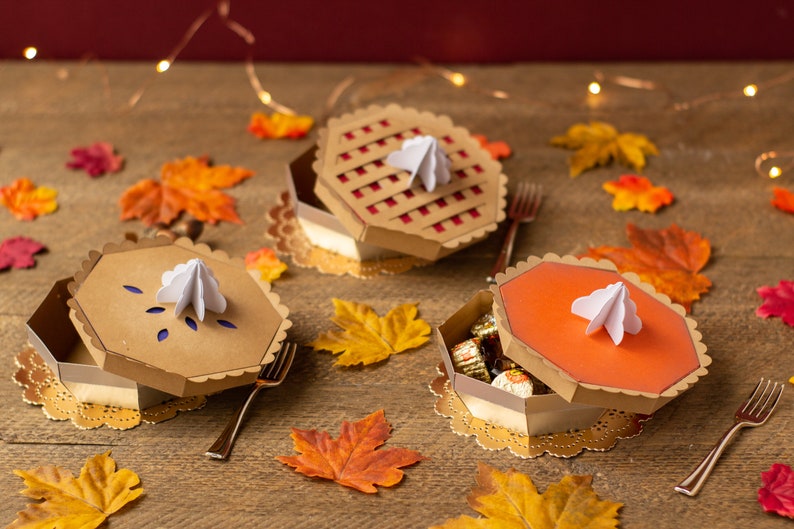 May include: Three pie-shaped boxes with lids, decorated with different pie fillings, on a wooden table with fall leaves. The boxes are open and filled with candy.