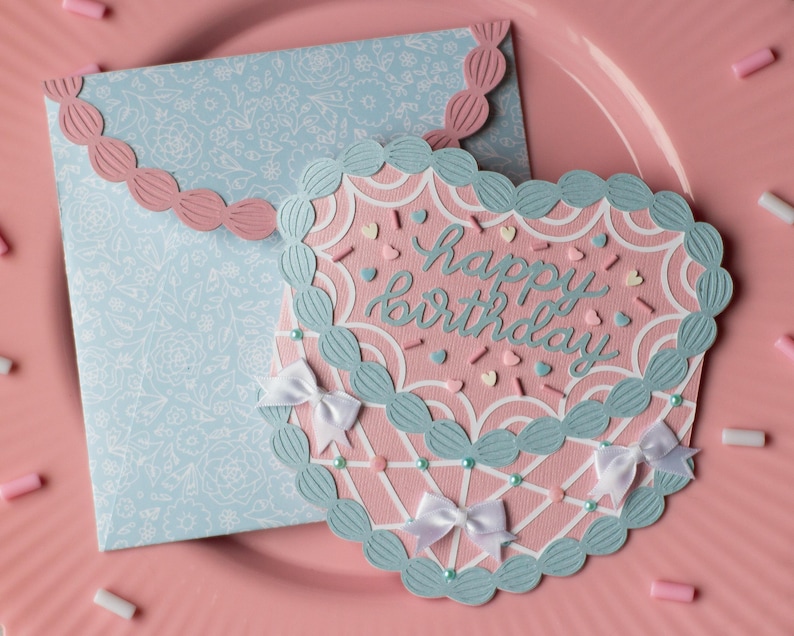 Pu&ograve; includere: Un biglietto a strati rosa e blu con un design a forma di cuore di torta. Il biglietto ha un bordo smerlato e dice "buon compleanno" in scrittura blu.