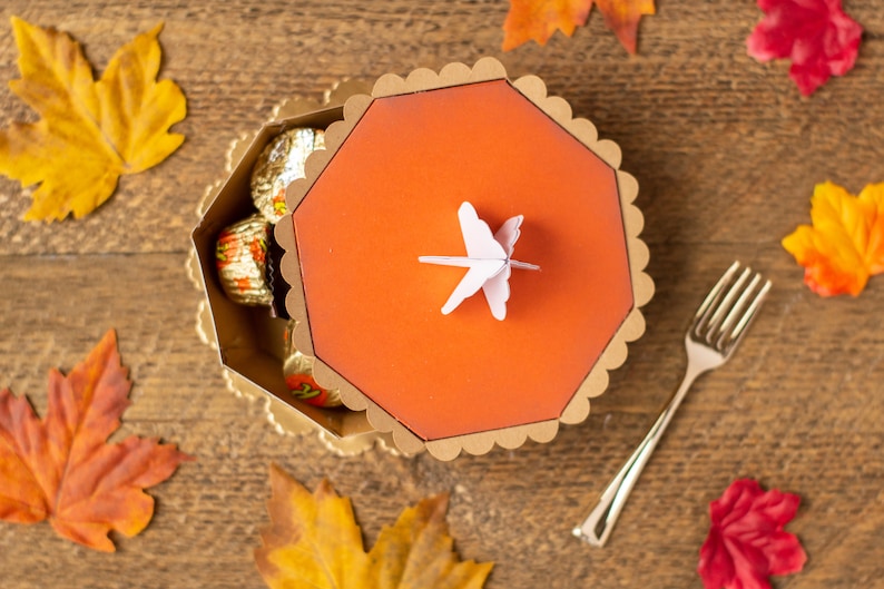 May include: A gold and orange octagonal box with a scalloped edge and a white paper butterfly on top. The box is filled with chocolate candies wrapped in gold foil.