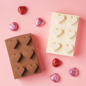 May include: Two boxes of chocolate hearts, one brown and one white, with a pink background. The boxes are filled with heart-shaped chocolates. There are also several loose chocolate hearts scattered around the boxes.