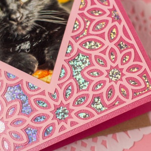 May include: A pink card with a cut-out design featuring a repeating floral pattern. The card has a photo of a black cat peeking through the cut-out design.