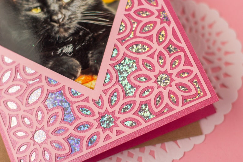 May include: A pink card with a cut-out design featuring a repeating floral pattern. The card has a photo of a black cat peeking through the cut-out design.