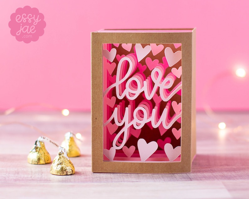 Puede incluir: Una caja de papel marr&oacute;n con una ventana que muestra un dise&ntilde;o de papel cortado en capas de color rosa y blanco con corazones y las palabras "love you".