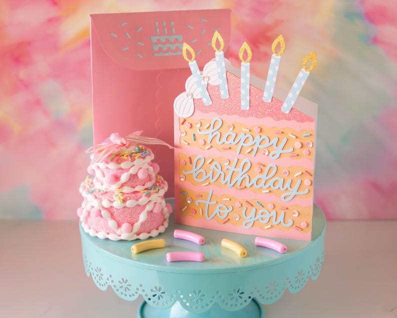 Może przedstawiać: R&oacute;żowa kartka urodzinowa w kształcie kawałka tortu z napisem "happy birthday to you" w jasnoniebieskim kolorze. Kartka ma błyszczące świeczki i posypkę. Małe r&oacute;żowe ciasto i cukierki stoją na niebieskim stojaku na ciasto.