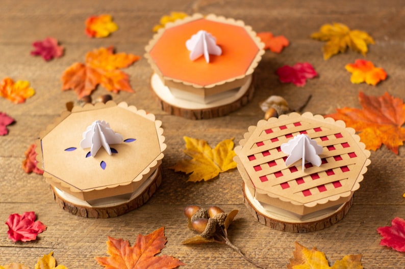 May include: Three paper pie boxes with different designs. The boxes are brown with red, orange, and blue accents. Each box has a white paper swirl on top. The boxes are sitting on a wooden surface with fall leaves scattered around them.
