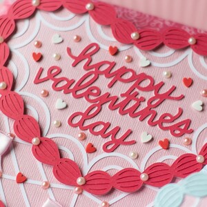 Pu&ograve; includere: Un biglietto rosa con un ritaglio a forma di cuore bianco e un bordo rosso. Il biglietto dice "Happy Valentine's Day" in lettere rosse. Il biglietto &egrave; decorato con cuori bianchi e rosa e perle bianche.