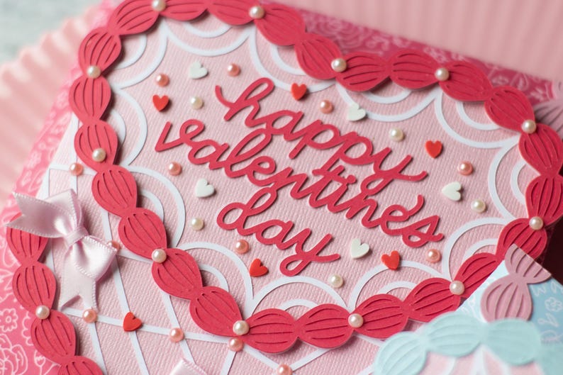 Pu&ograve; includere: Un biglietto rosa con un ritaglio a forma di cuore bianco e un bordo rosso. Il biglietto dice "Happy Valentine's Day" in lettere rosse. Il biglietto &egrave; decorato con cuori bianchi e rosa e perle bianche.