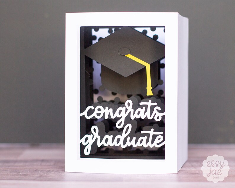 Puede incluir: Tarjeta blanca con un patr&oacute;n de lunares negros y blancos dentro de una ventana. Un gorro de graduaci&oacute;n negro con una borla amarilla est&aacute; dentro de la ventana. Las palabras "congrats graduate" est&aacute;n escritas en cursiva blanca en la tarjeta.