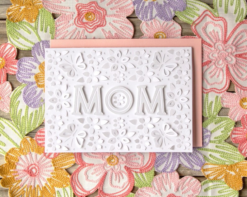 May include: White card with a pink backing, featuring a white paper cut-out design of flowers and the word "MOM". The card is surrounded by a floral fabric.