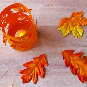 May include: Orange candle holder with a leaf design, surrounded by three orange and yellow leaves and a string of white lights on a wood background.