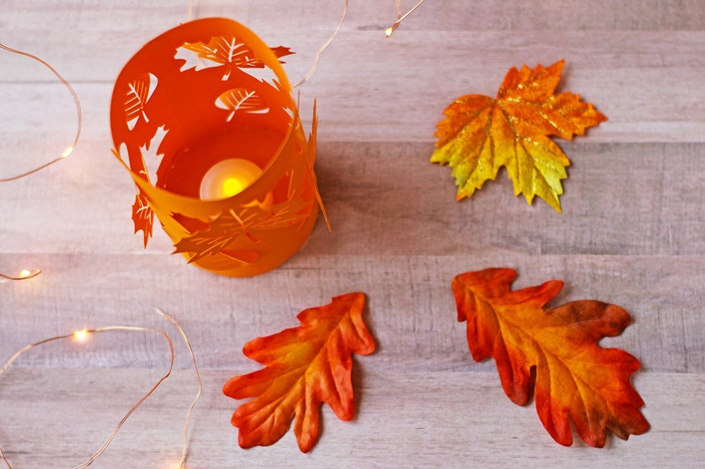 May include: Orange candle holder with a leaf design, surrounded by three orange and yellow leaves and a string of white lights on a wood background.
