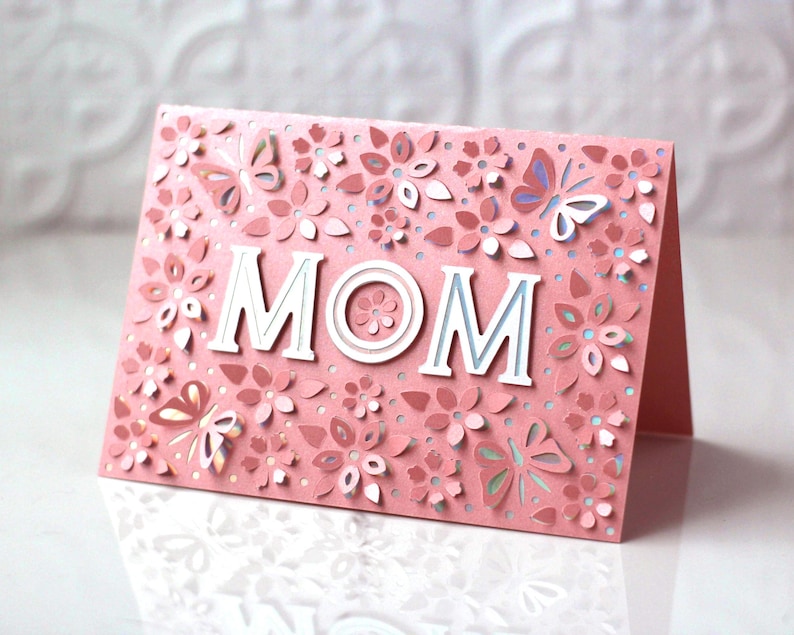 May include: Pink card with a delicate floral design and the word "MOM" in white letters. The card has a 3D effect with the flowers and butterflies raised.
