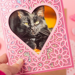 May include: A pink card with a heart-shaped cutout featuring a black cat. The card has a glittery, floral design.