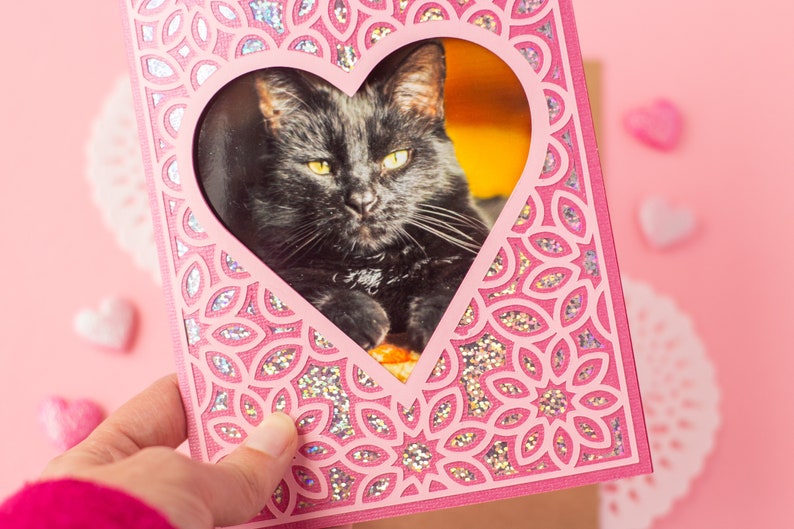 May include: A pink card with a heart-shaped cutout featuring a black cat. The card has a glittery, floral design.
