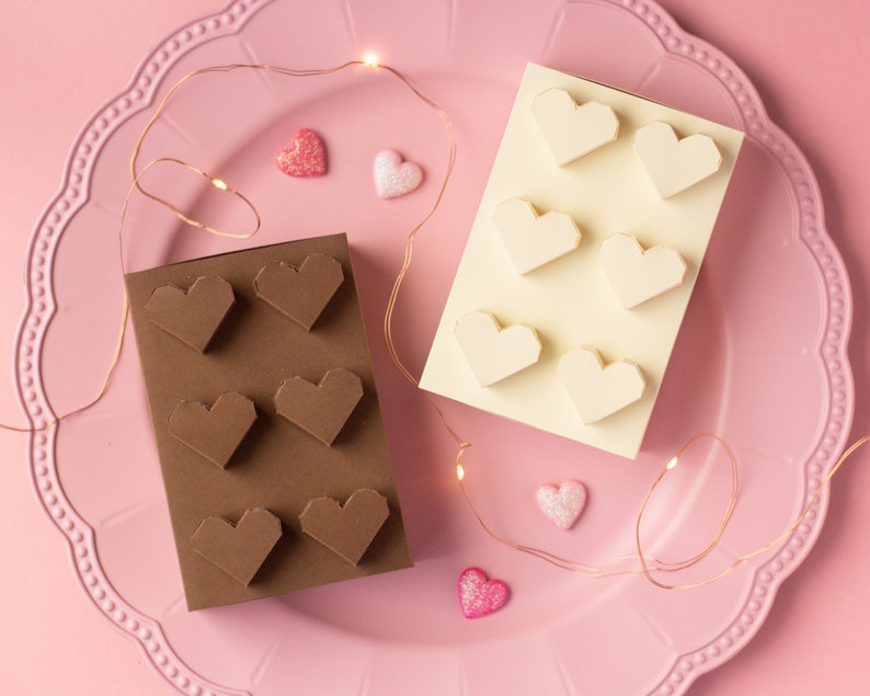 May include: Two white and brown paper cards with heart-shaped cutouts. The cards are on a pink plate with a decorative edge. The plate is surrounded by a string of fairy lights.