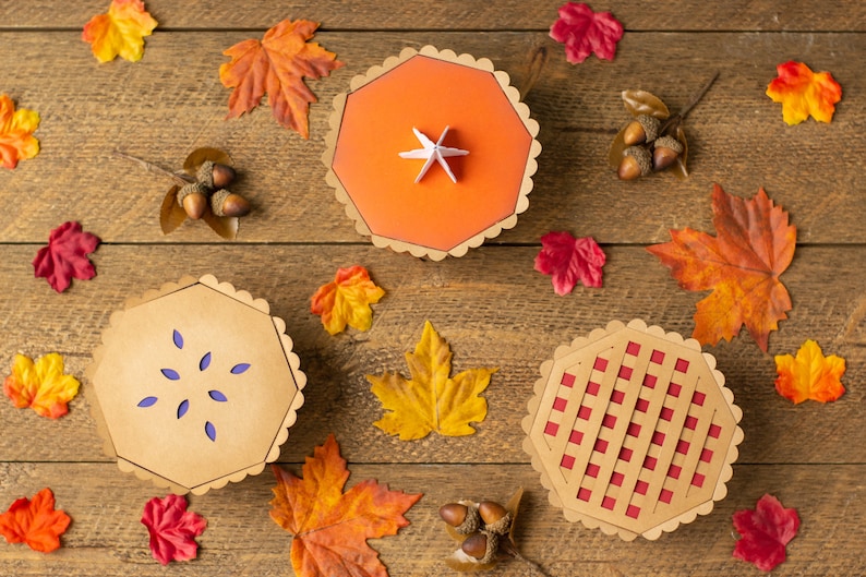 May include: Three paper pie boxes with different designs. The boxes are brown with scalloped edges. One box has a red and white checkered design, one has a solid orange design with a white star, and one has a purple design with a starburst pattern.