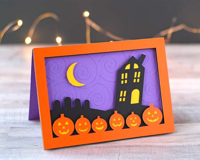 May include: A Halloween card with a purple background and a black silhouette of a house with a yellow window. The card also features a crescent moon and five orange pumpkins with black faces.
