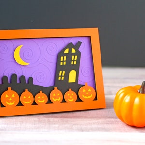 May include: A Halloween card with a purple background and a black silhouette of a house with yellow windows. The house is surrounded by a row of orange pumpkins with black faces. There is a crescent moon in the sky.