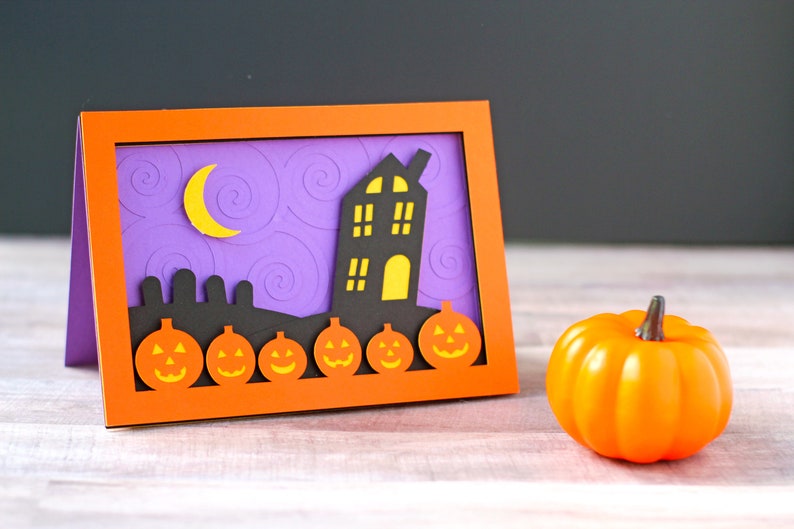 May include: A Halloween card with a purple background and a black silhouette of a house with yellow windows. The house is surrounded by a row of orange pumpkins with black faces. There is a crescent moon in the sky.