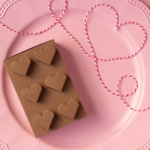 May include: A pink plate with a red and white string tied in a heart shape. On the plate is a chocolate bar shaped like a brick with heart-shaped cutouts.