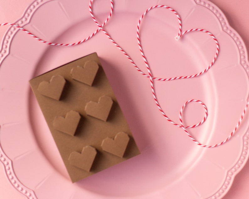 May include: A pink plate with a red and white string tied in a heart shape. On the plate is a chocolate bar shaped like a brick with heart-shaped cutouts.