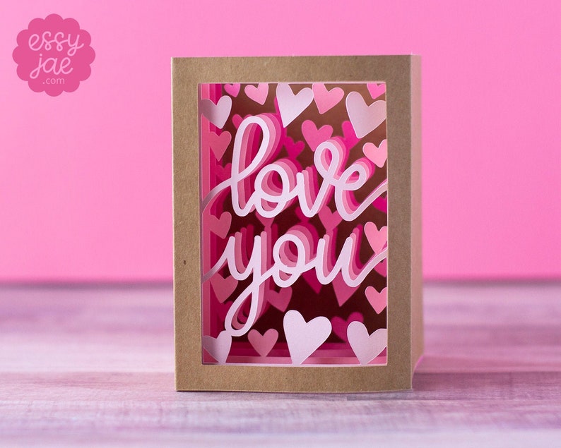 Puede incluir: Una tarjeta marr&oacute;n con una ventana recortada. La ventana est&aacute; llena de corazones de papel rosa y blanco y las palabras "love you" en papel rosa y blanco.