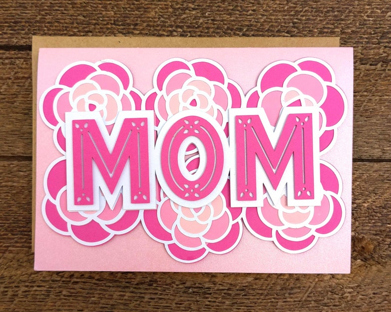 May include: A pink greeting card with the word "MOM" in bold, hot pink letters. The letters are outlined in white and set against a backdrop of layered pink and white floral designs. The card is on a light pink background.