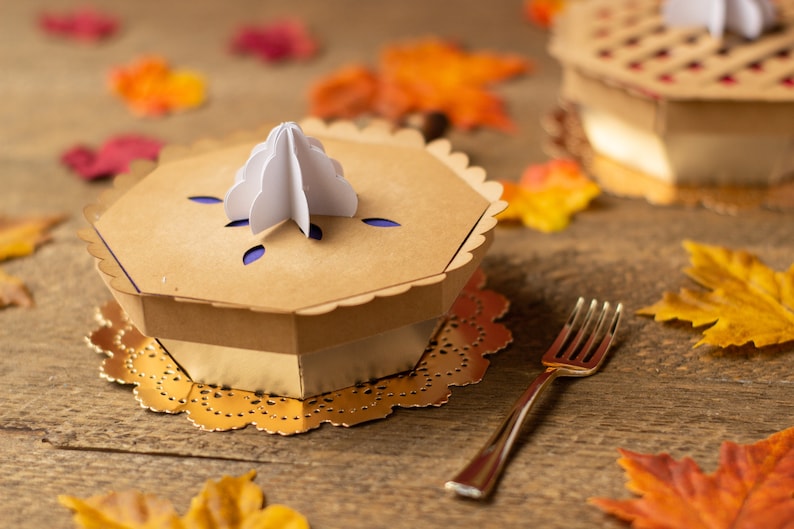 May include: A brown cardboard pie box with a white paper pie crust topper. The box is sitting on a gold doily on a wooden surface. There are fall leaves scattered around the box.