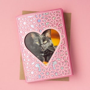 May include: Pink card with a heart-shaped window featuring a black cat. The card has a glittery, patterned design.