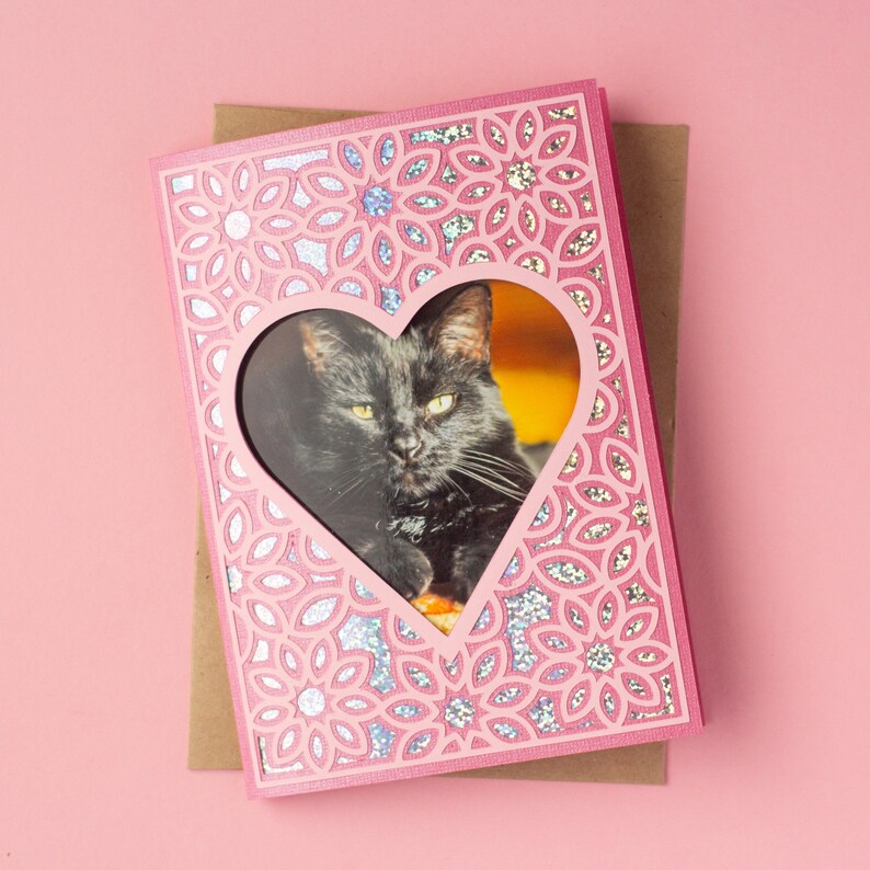 May include: Pink card with a heart-shaped window featuring a black cat. The card has a glittery, patterned design.