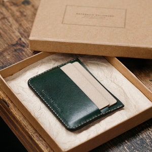 May include: A dark green leather card holder with a brown cardboard box. The card holder has a stitched edge and is made of high-quality leather.