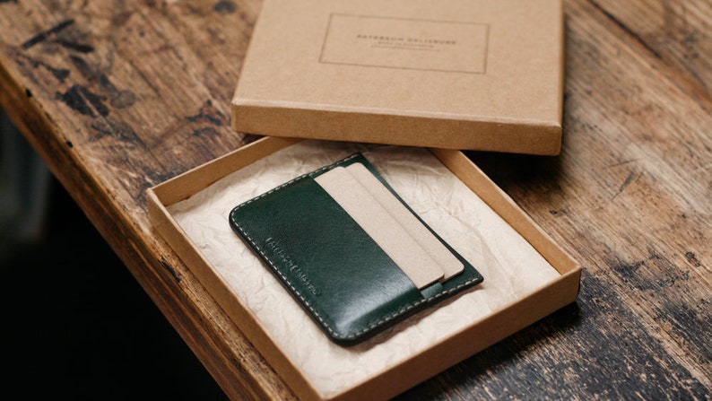 May include: A dark green leather card holder with a brown cardboard box. The card holder has a stitched edge and is made of high-quality leather.
