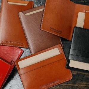 May include: A collection of six leather wallets in various colors, including red, brown, and black. The wallets are made of high-quality leather and have a simple, minimalist design. Each wallet features the text "Leather Society" embossed on the front.