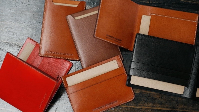 May include: A collection of six leather wallets in various colors, including red, brown, and black. The wallets are made of high-quality leather and have a simple, minimalist design. Each wallet features the text "Leather Society" embossed on the front.