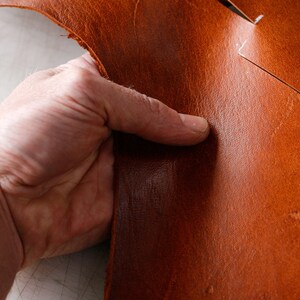 May include: A close-up of a hand holding a piece of brown leather. The leather has white markings on it, indicating where it should be cut.