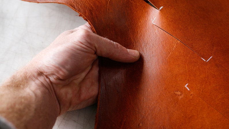 May include: A close-up of a hand holding a piece of brown leather. The leather has white markings on it, indicating where it should be cut.