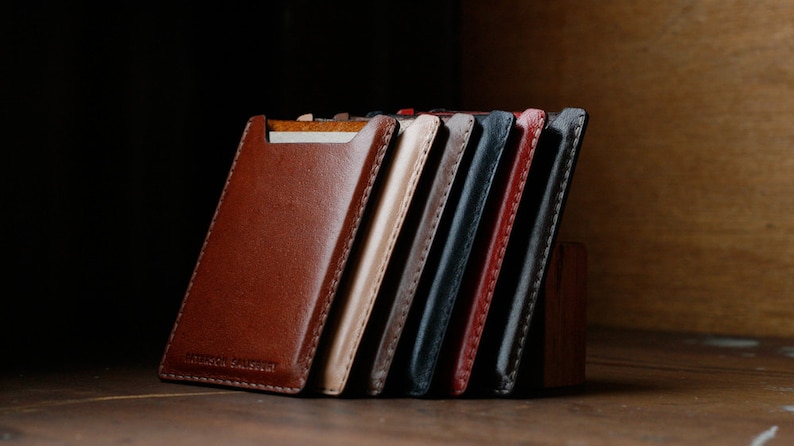 May include: A set of eight leather card holders in various colors, including brown, tan, gray, black, and red. The holders are stacked on top of each other and have a stitched edge.