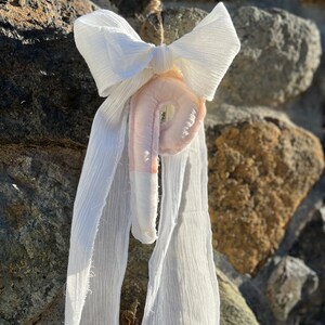 May include: A handmade, off-white fabric ornament with a bow and flowing ribbons. The ornament has a light pink center with a circular opening. It is suspended by a natural twine string against a stone wall.