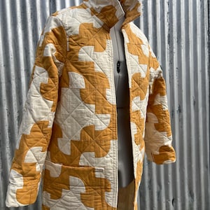 May include: A long, quilted coat in a cream and mustard yellow color scheme. The coat features a geometric pattern and a high collar. The coat is displayed on a mannequin. The coat has two pockets and a zipper closure.
