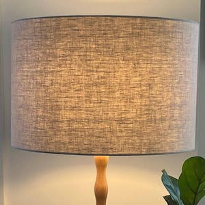 May include: A cylindrical lampshade in light grey fabric with a dark grey trim, mounted on a wooden lamp base. The lamp is lit, emitting a warm light. A green plant with large leaves is in the foreground.