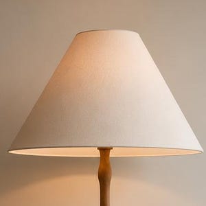 May include: A floor lamp featuring a slender, turned wooden stand with a natural light brown finish and a simple, conical off-white fabric lampshade that emits a warm, soft glow from the light source positioned inside the shade.
